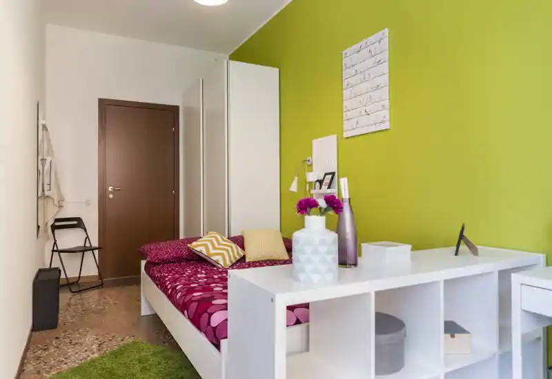 Bright, modern single bedroom with a patterned pink bedspread, decorative cushions and a green accent wall. White shelving and wardrobe provide storage while a small desk area and accessories create a cozy, well-styled space.