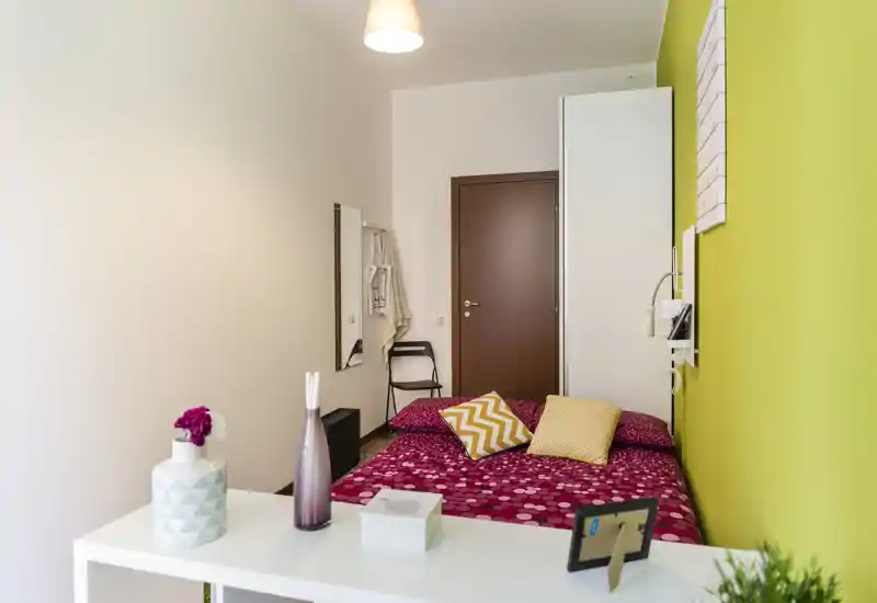 Compact, well-staged bedroom with a patterned double bed, colorful cushions and a bright green accent wall; simple storage and decorative vases complete the cozy setup.
