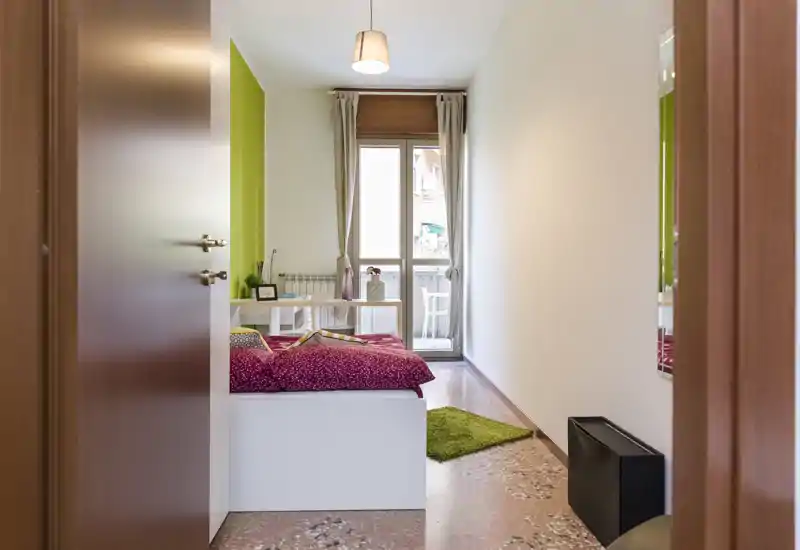 Bright, modern bedroom with a double bed framed by a green accent wall, a desk by the window and natural light from the balcony door.