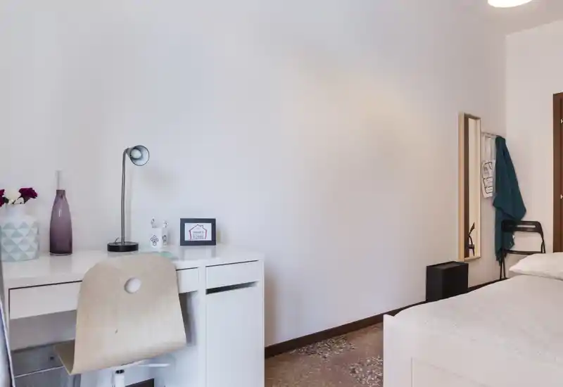 Minimal, well-lit bedroom corner with a white bed and a small desk. The space feels tidy and functional with simple decorative touches and a standing mirror by the door.