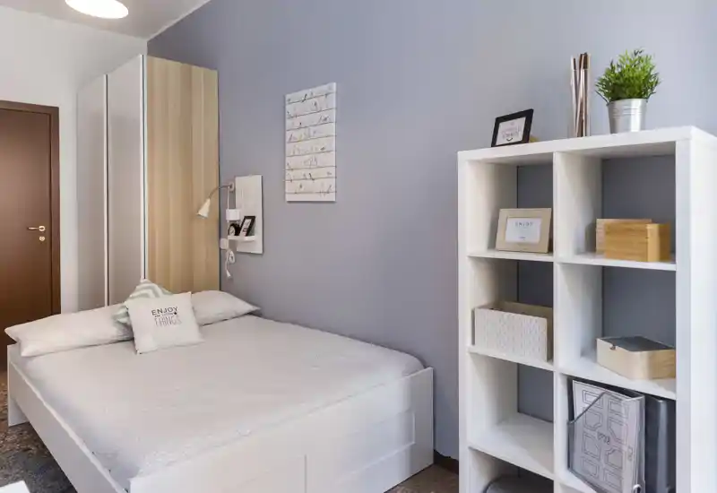 Camera da letto luminosa e ben allestita con letto bianco, parete grigio-blu e scaffalatura ordinata; perfetta per foto in affitto che mostrano un'area notte moderna e rilassante.