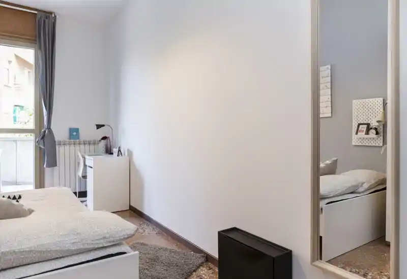 Bright, modern bedroom with a white bed, soft textiles and a desk by the window. Neutral décor, natural light and a full-length mirror that adds depth.
