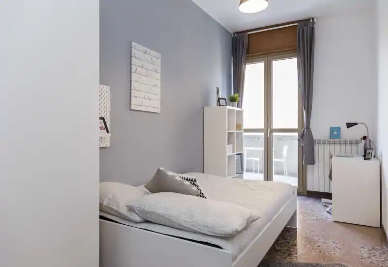 Bright, modern bedroom with a double bed in the foreground, neutral linens and a balcony door providing natural light. Compact shelving and a small desk create a tidy, functional space ideal for single or couple stays.