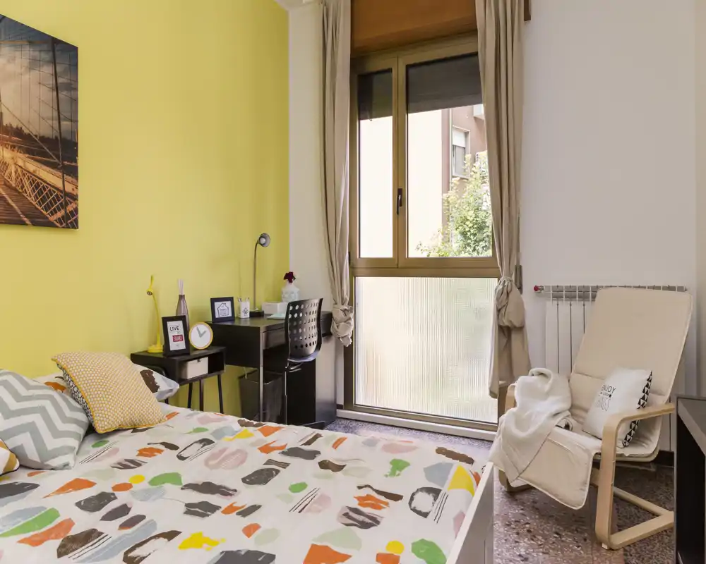 Chambre lumineuse et soignée avec une couette colorée, coussins décoratifs, un petit bureau près de la fenêtre et un fauteuil confortable — idéale pour une personne ou un étudiant.