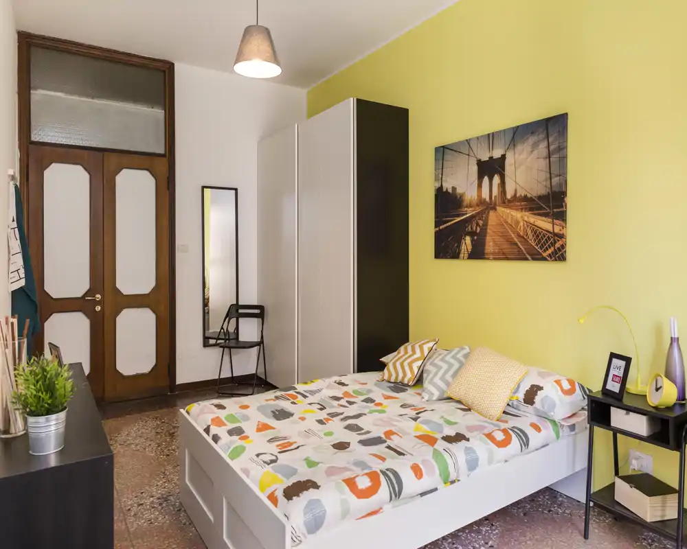 Chambre lumineuse et chaleureuse avec un lit double, une couette à motifs, une table de chevet et une armoire. Mur jaune et tableau apportent une atmosphère accueillante.
