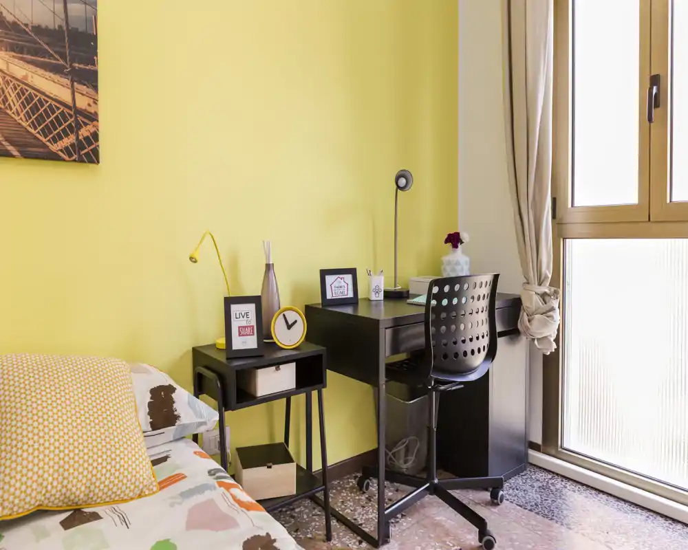 Coin chambre lumineux et compact avec lit et petit espace bureau : literie à motifs, table de chevet avec lampe et horloge, et un bureau en bois sombre près de la fenêtre pour un coin travail lumineux.