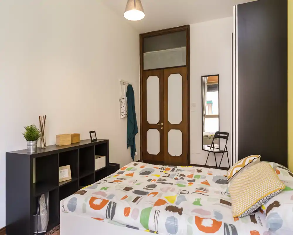 Chambre chaleureuse et lumineuse avec une couette à motifs, des coussins décoratifs et une étagère de rangement simple. On note une grande armoire, un miroir pleine longueur et une porte en bois élégante.