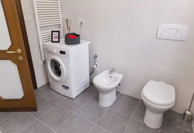 Compact, well-lit bathroom with a toilet, bidet and a front-loading washing machine; neutral tiles and clean modern fixtures.