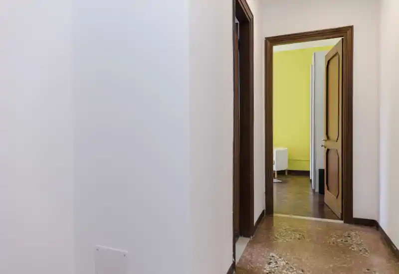 Simple, well-lit entrance hallway with a visible wooden doorway leading into a bright room with yellow walls; polished patterned floor and clean white walls create a neutral transitional space.