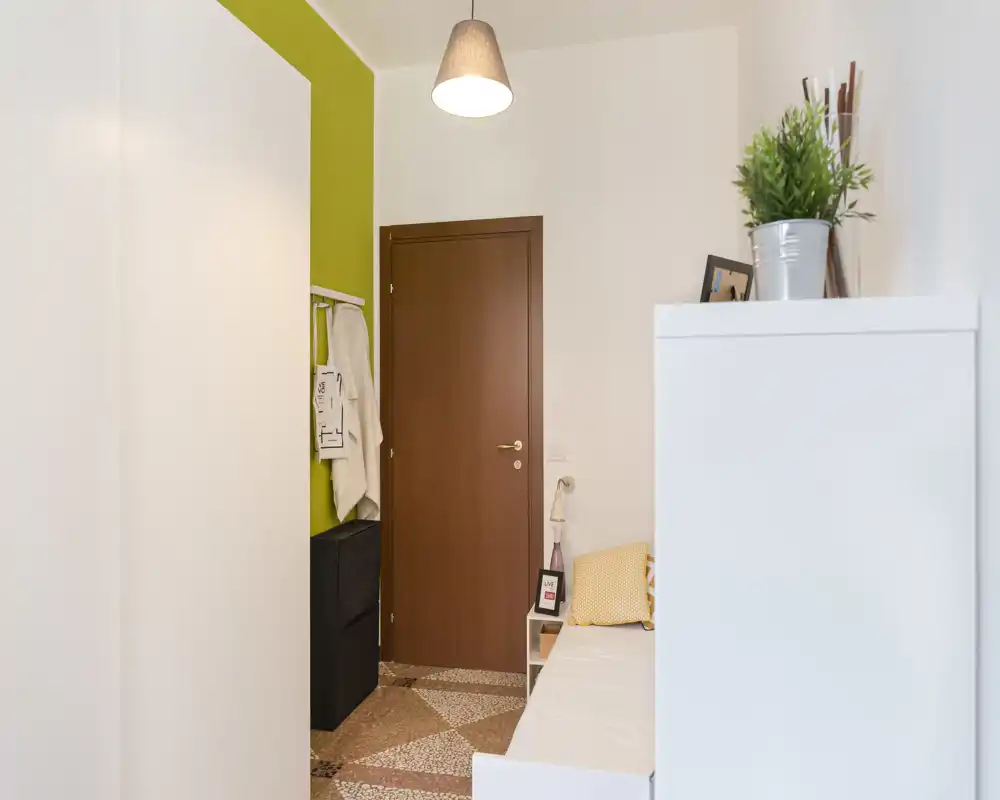 Compact bedroom/entry nook with a single bed/bench, warm pendant light and bright white walls accented in green; tidy, modern and functional for a small urban apartment.