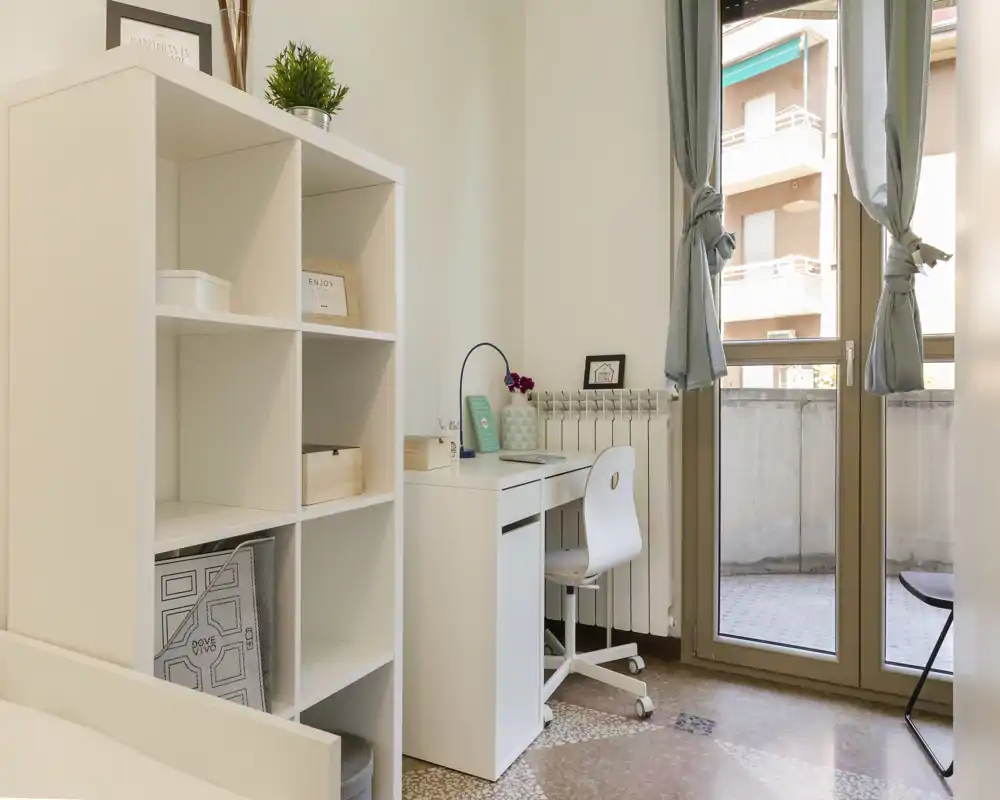 Bright, minimal bedroom corner featuring a white desk and shelving with a small visible bed frame at the edge; natural light from the balcony door creates a fresh, airy feel.