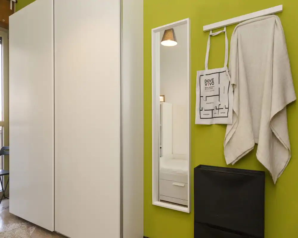 Small bedroom corner with a white wardrobe, a tall mirror reflecting a bedside area and soft textiles hung on hooks; clean, modern styling with warm light.