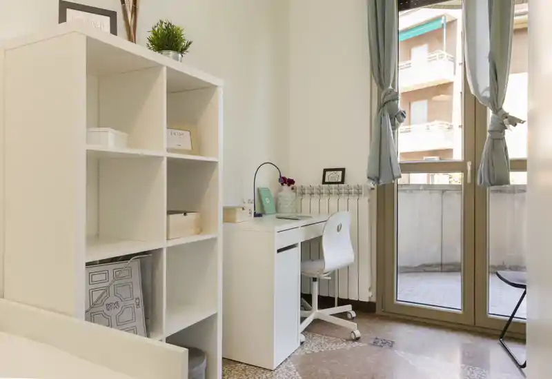Bright, minimal bedroom corner featuring a white desk and shelving with a small visible bed frame at the edge; natural light from the balcony door creates a fresh, airy feel.
