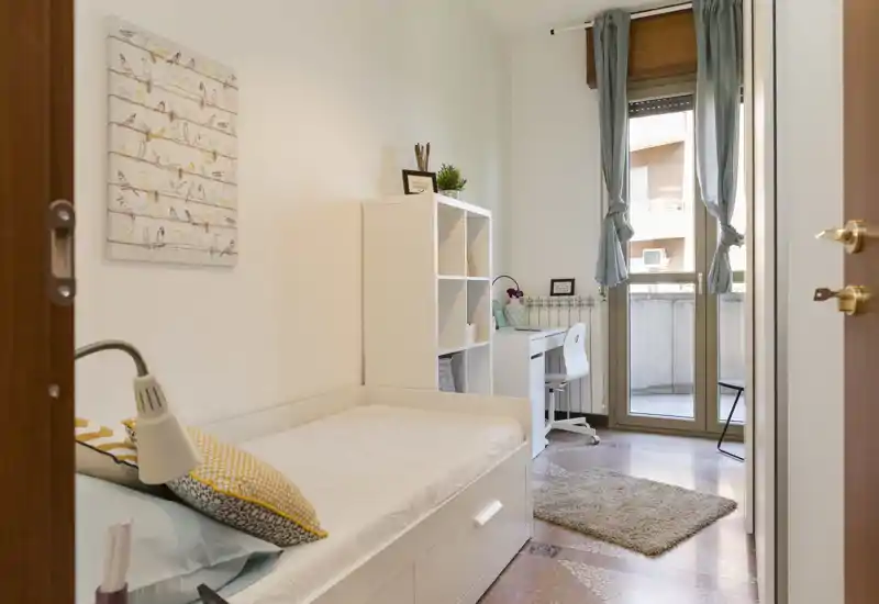 Bright, well-staged single bedroom with a neatly made bed, shelving and a desk by the balcony door letting in natural light — ideal for a student or single occupant.