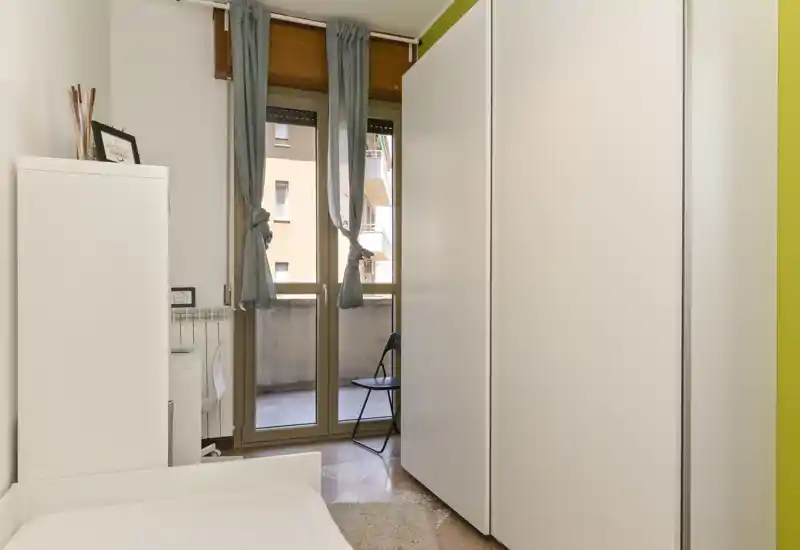 Compact bedroom with a visible single bed in the foreground, a large white wardrobe, and French-door windows letting in natural light; simple, tidy styling suitable for a rental listing.
