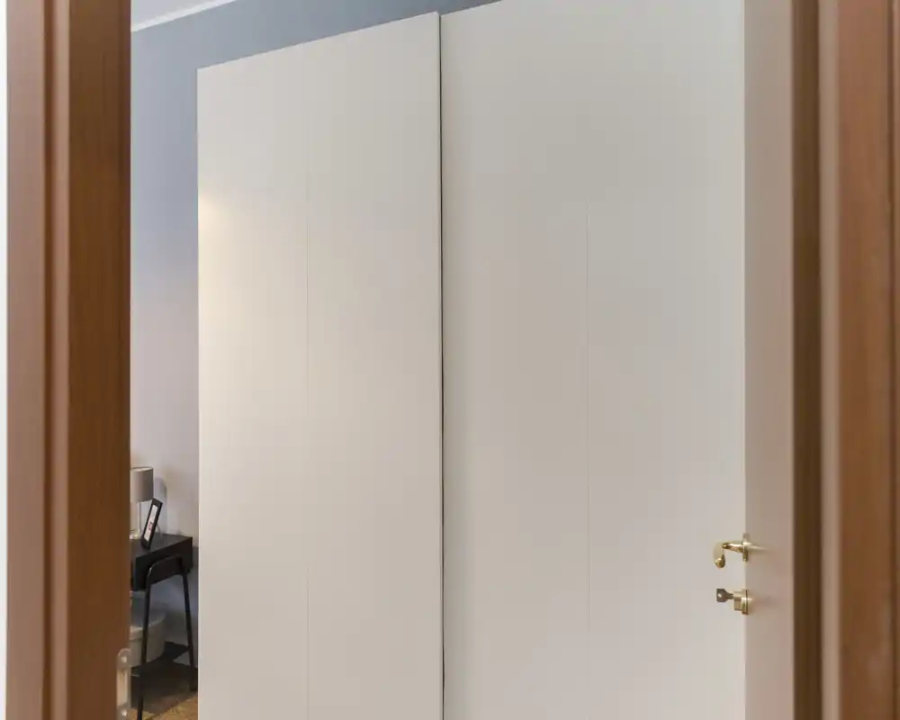 Vista attraverso una porta che mostra ante bianche di un armadio a muro e un parziale scorcio di un comodino con lampada a sinistra; la funzione della stanza non è chiaramente mostrata.