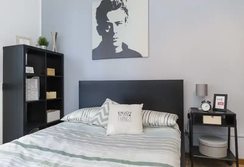 Camera da letto luminosa e ordinata con letto matrimoniale, cuscini decorativi e comodino con lampada e sveglia. Palette neutra e scaffalatura semplice creano un’atmosfera moderna e rilassante.