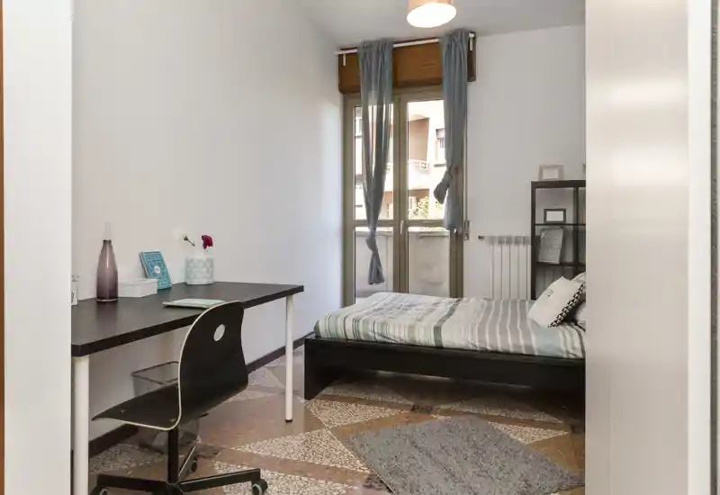 Camera da letto luminosa e ordinata con letto matrimoniale, scrivania semplice e luce naturale dalla porta del balcone — ideale per studenti o single.