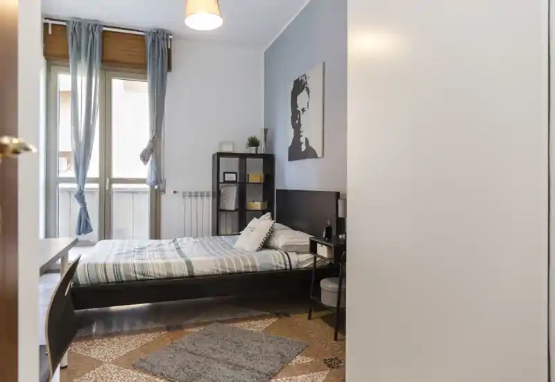 Camera da letto luminosa e ben arredata con letto matrimoniale, biancheria a righe, comodino e scaffale; ampia finestra che garantisce luce naturale e un’atmosfera gradevole.