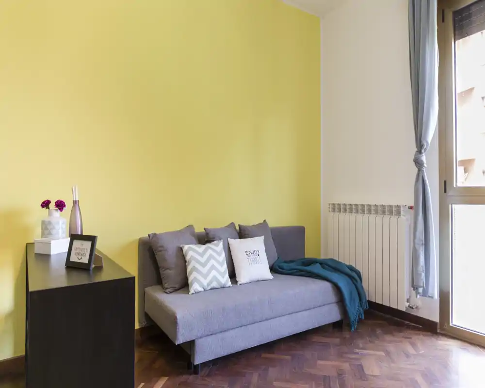 Bright, modern living area with a compact grey sofa, decorative cushions and a teal throw against a warm yellow accent wall. Natural light enters from a large window, creating a welcoming, well-styled small lounge.