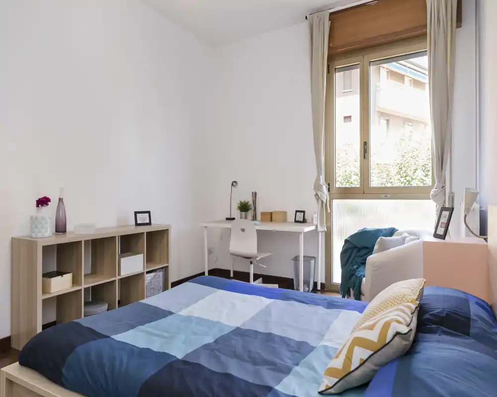 Bright, tidy bedroom with a double bed dressed in blue patchwork bedding, a desk by the window and light wood shelving — ideal for comfortable stays and remote work.