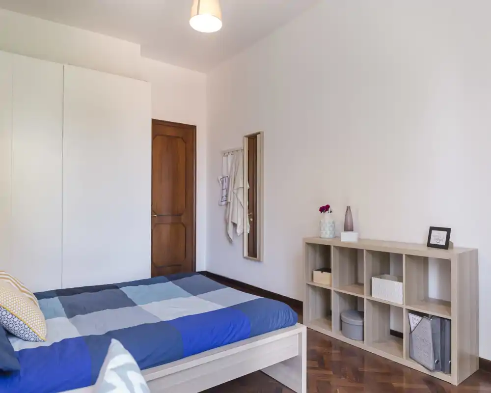 Bright, neatly staged bedroom with a double bed dressed in blue patchwork linens, a large wardrobe, and open shelving—ideal for rental listings emphasizing clean, modern comfort.