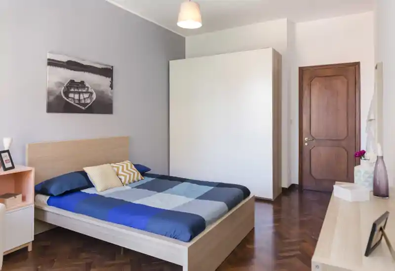 Bright, modern bedroom with a full wooden bed, patterned cushions and a large white wardrobe; parquet flooring and soft natural light create a welcoming atmosphere ideal for rental photos.