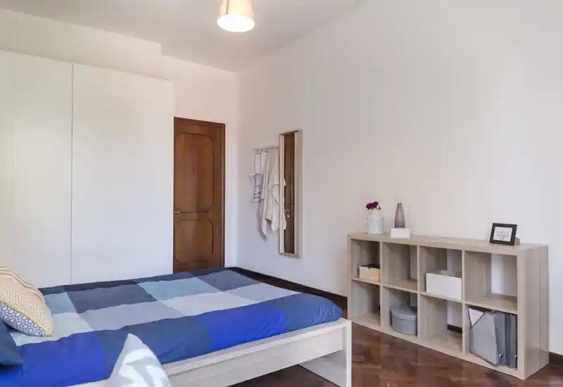 Bright, neatly staged bedroom with a double bed featuring blue patchwork bedding, a large wardrobe, and open shelving — ideal for rental listings highlighting clean lines and natural light.