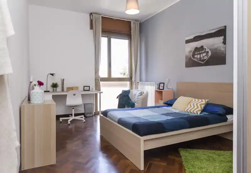 Bright, well-staged bedroom with a double bed, work desk by the window and calm blue-gray accent wall. Natural light fills the room and warm wooden flooring adds a cozy touch.