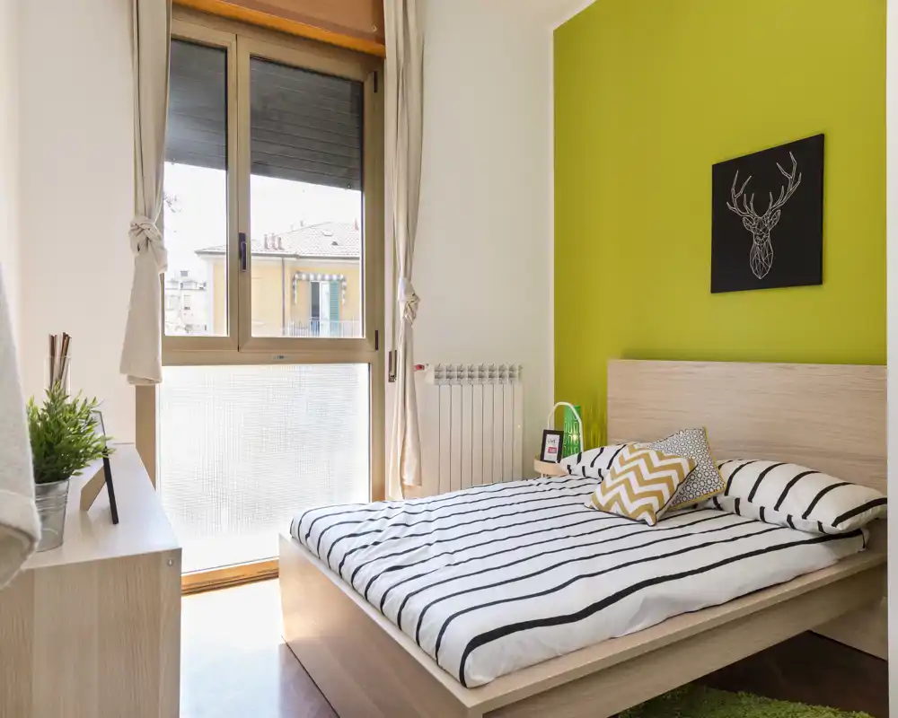 Camera da letto moderna e luminosa con letto su pedana in legno, biancheria a righe e una finestra ampia che illumina l'ambiente. La parete verde e i cuscini decorativi creano un tocco accogliente perfetto per affitti brevi.
