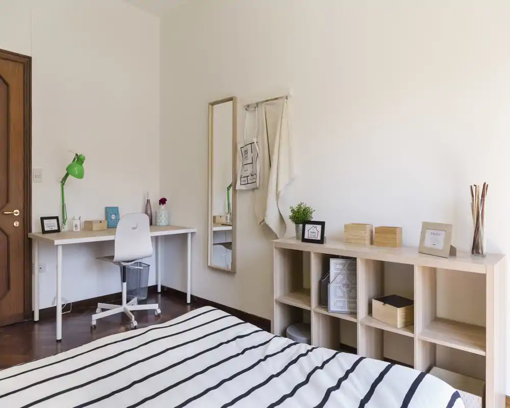 Camera da letto luminosa e minimal con copriletto a righe in primo piano, scrivania ordinata con lampada verde, specchio a figura intera e scaffalatura aperta—spaziosa, ben illuminata e dallo stile semplice.