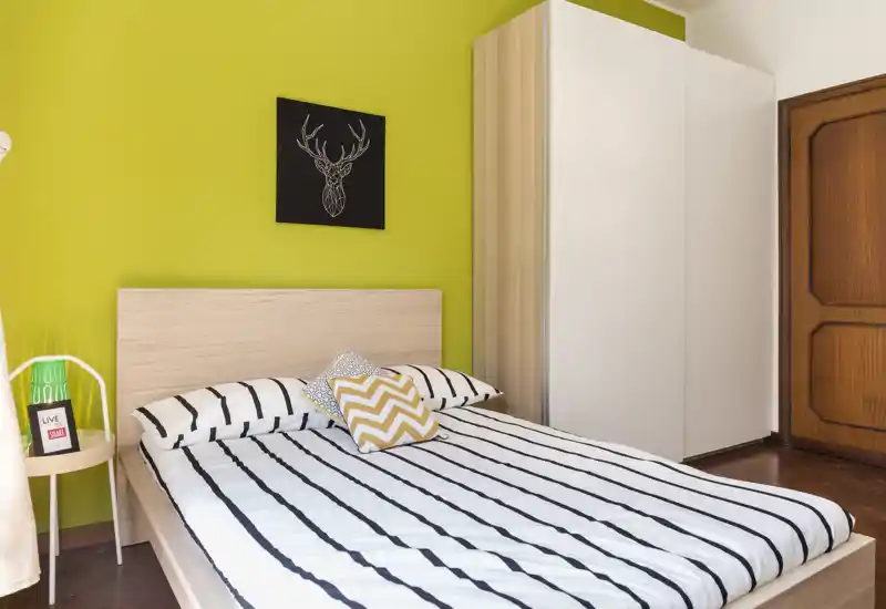 Camera da letto moderna e luminosa con letto matrimoniale a righe, cuscini decorativi e testiera in legno chiaro su una parete verde brillante. Un armadio bianco e una porta in legno completano l’aspetto ordinato.