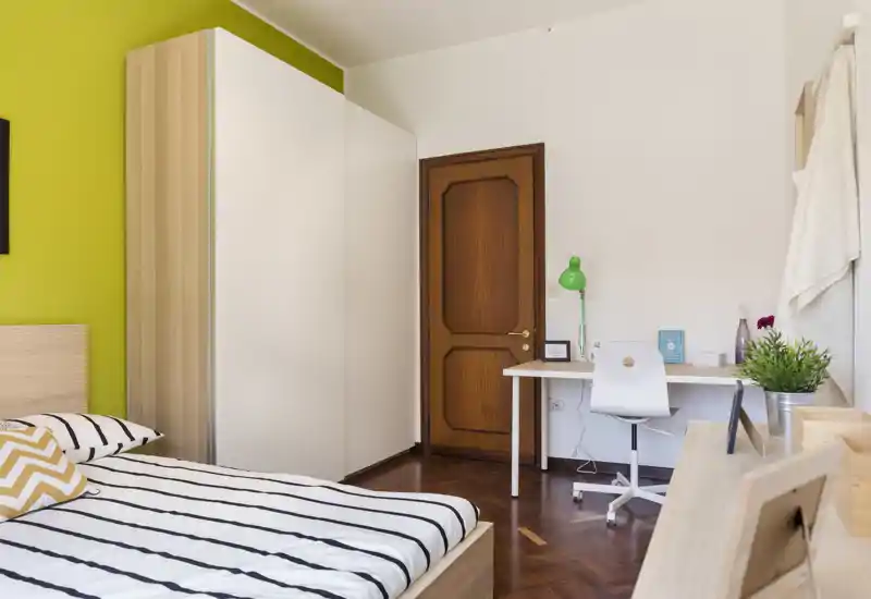 Camera da letto luminosa e ben arredata con letto matrimoniale a righe, grande armadio e una scrivania ordinata — perfetta per riposo e lavoro.