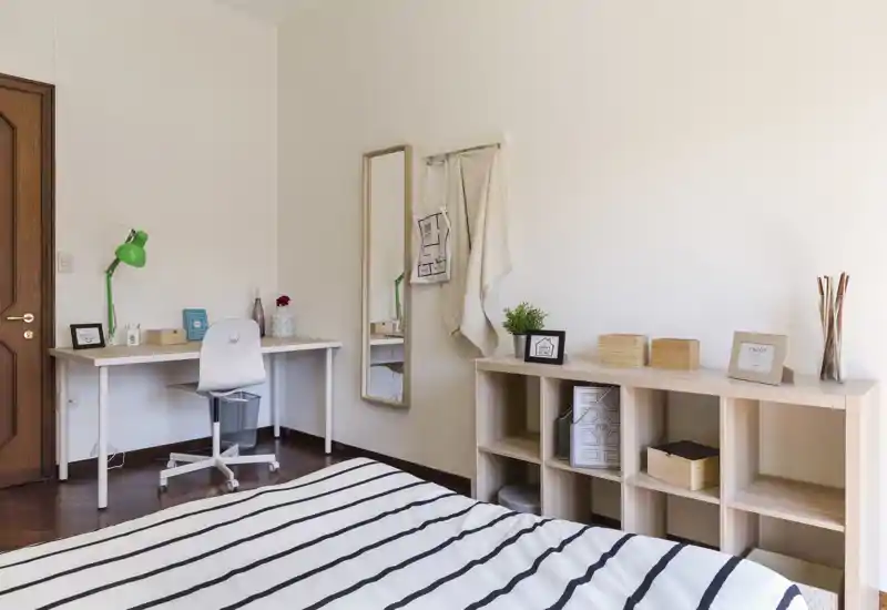 Bright, minimal bedroom with a striped bed in the foreground, a tidy desk with a green lamp, full-length mirror and open shelving—clean, well-lit and simply styled.