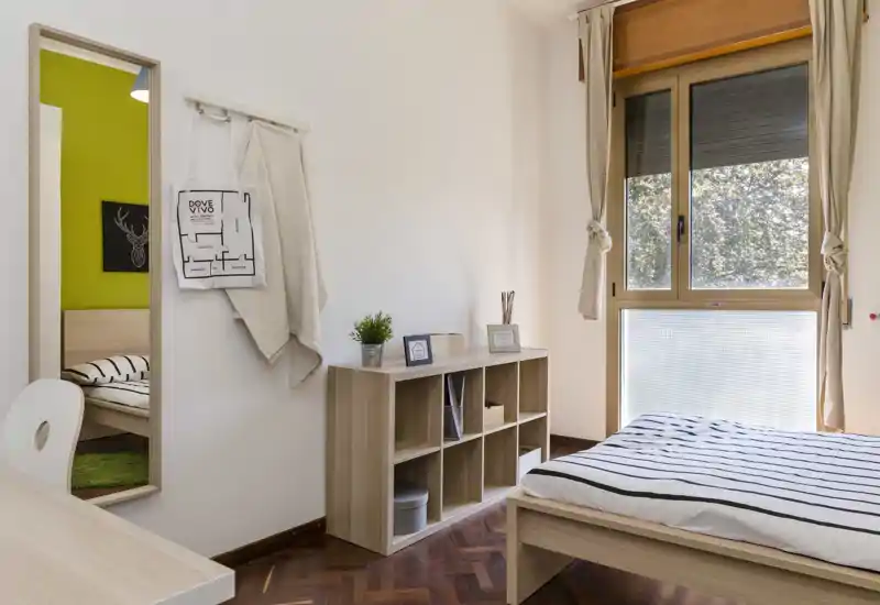 Camera da letto luminosa e curata con letto a righe, mobile contenitore a cubi in legno e grande finestra che lascia entrare luce naturale—perfetta per annunci.