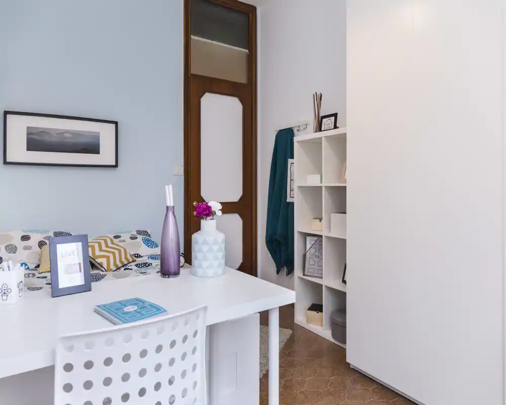 Bright, neatly styled bedroom corner with a white desk in the foreground, patterned pillows and a bed visible against a pale blue wall, plus shelving and decorative accents.