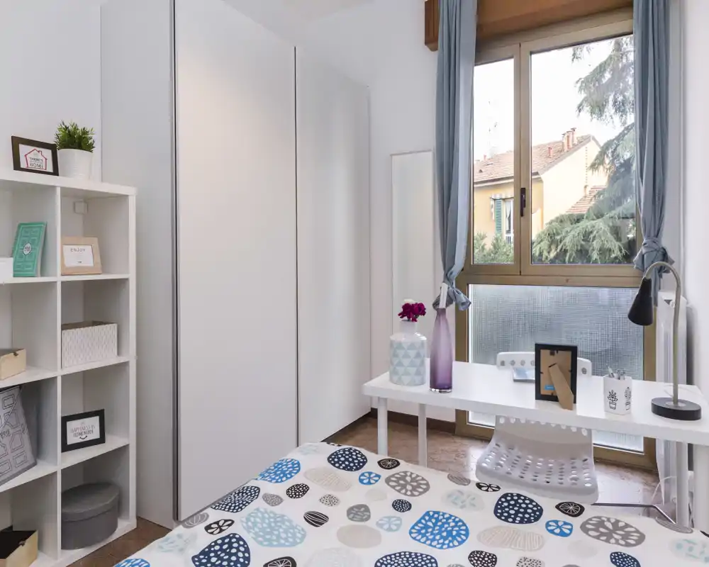 Sunny, well-staged bedroom with a patterned bedspread, white wardrobe and a desk by the window offering natural light and a pleasant view.