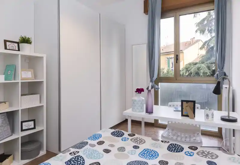Sunny, well-staged bedroom with a patterned bedspread, white wardrobe and a desk by the window offering natural light and a pleasant view.