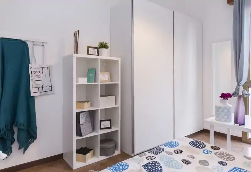 Bright, modern bedroom corner with patterned bedding, a white wardrobe and open shelving; tasteful decorative items and natural light create a welcoming atmosphere.