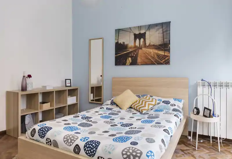 Camera da letto luminosa e ben arredata con cornice letto in legno, biancheria con motivo e cuscini decorativi. Una libreria bassa e uno specchio a figura intera aggiungono spazio contenitivo e profondità; un quadro e toni chiari creano un'atmosfera rilassante.