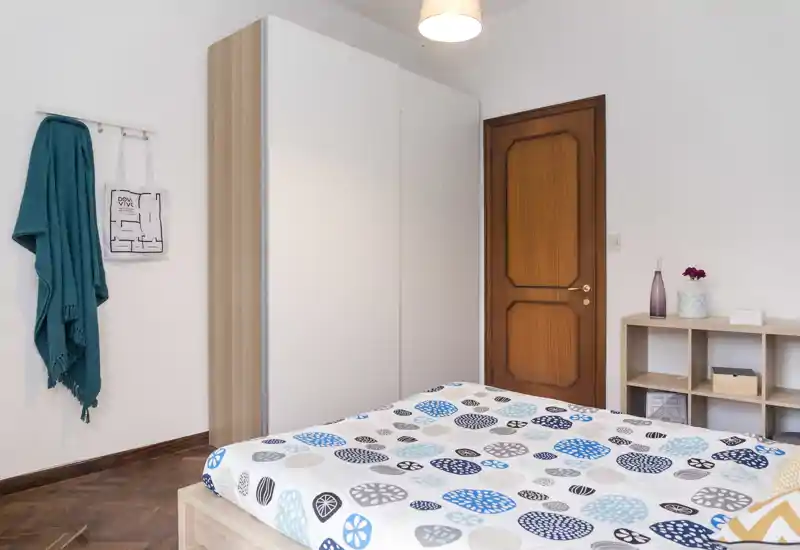 Camera da letto luminosa e dallo stile semplice con un letto matrimoniale a fantasia in primo piano, un ampio armadio bianco, porta in legno e piccolo scaffale — ordinata e accogliente.