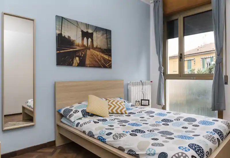 Chambre lumineuse et bien décorée avec lit double en bois, couette à motifs et coussins décoratifs. Grande fenêtre apportant une belle lumière naturelle et un miroir mural qui agrandit l'espace.