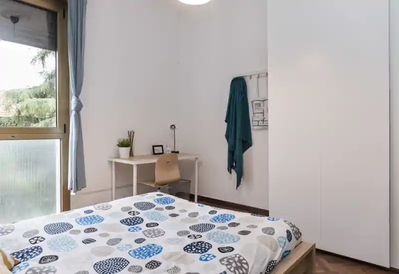 Camera da letto luminosa e ordinata con copriletto a fantasia, una piccola scrivania vicino alla finestra e un ampio armadio — confortevole e ben illuminata per una persona.