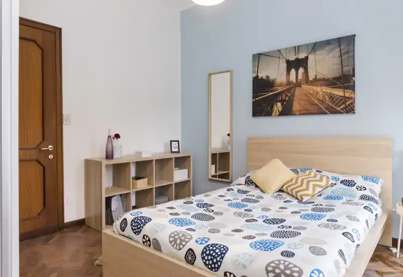 Camera da letto luminosa e ben allestita con letto in legno, biancheria a fantasia e cuscini decorativi; parete azzurra e quadro contribuiscono a un’atmosfera accogliente.