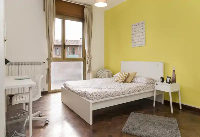 Dormitorio luminoso y ordenado con cama doble blanca frente a una pared amarilla, mesita, escritorio y una gran ventana que aporta luz natural.