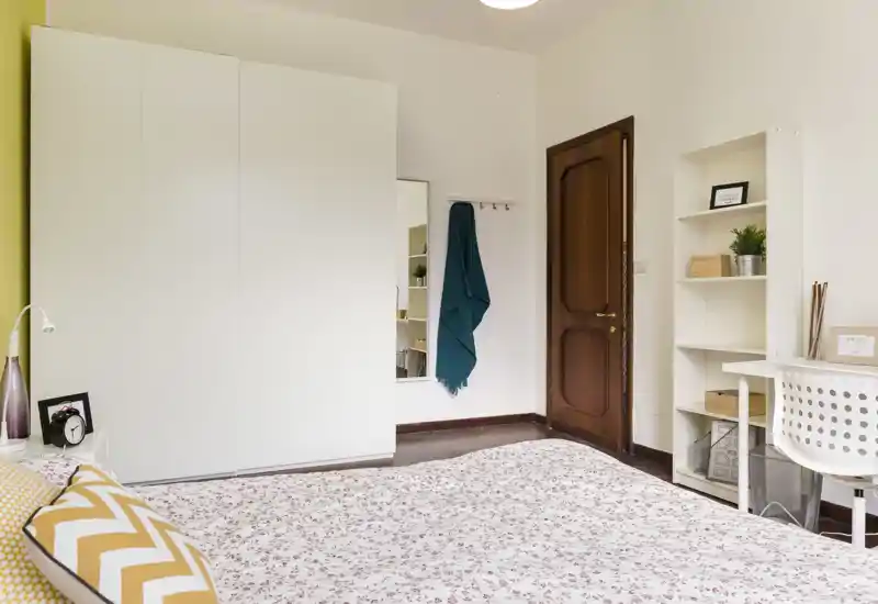 Dormitorio luminoso y ordenado con una cama grande con ropa de cama estampada, armario blanco, escritorio y estantería — una habitación acogedora y bien iluminada ideal para anuncios.