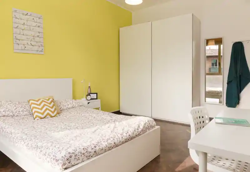 Dormitorio moderno y luminoso con cama doble, mobiliario blanco y un cojín chevron sobre edredón estampado; armario y escritorio completan el espacio acogedor y bien iluminado.