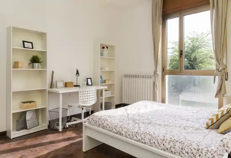 Dormitorio soleado y bien decorado con cama blanca, ropa de cama estampada y escritorio junto a la ventana. Suelo de madera clara y estanterías que crean un ambiente cálido y luminoso, perfecto para alquileres.