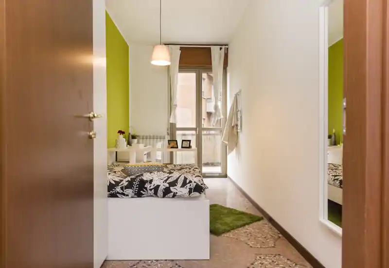 Camera singola luminosa e moderna con biancheria a fantasia, parete accent verde e finestra con accesso a un piccolo balcone. La stanza è ordinata e arredata con una mensola, una lampada sospesa e un tappeto che la rendono accogliente.