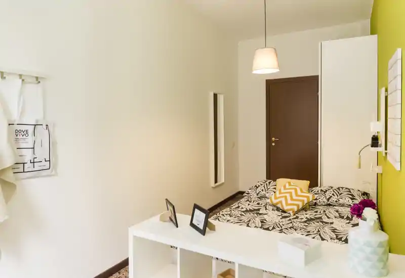 Camera da letto luminosa e ben arredata con letto a fantasia, cuscini decorativi e un piccolo divisorio a scaffale che separa l'area notte; lampada sospesa calda e parete verde accentuano un'atmosfera accogliente e moderna.